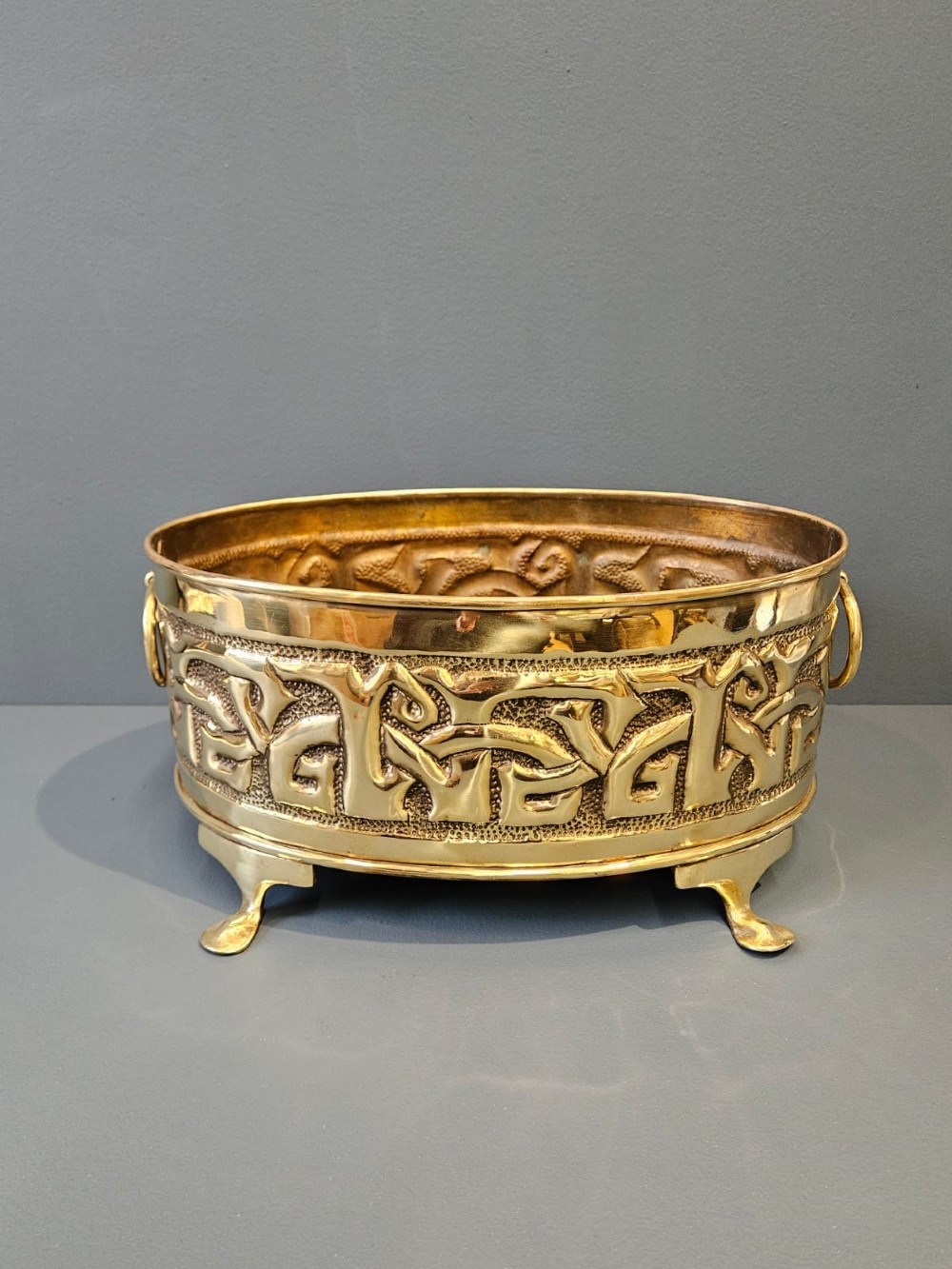 An oval polished brass planter featuring embossed Glasgow School Celtic knotwork on a stippled background, raised on short bracket feet with side handles. View II