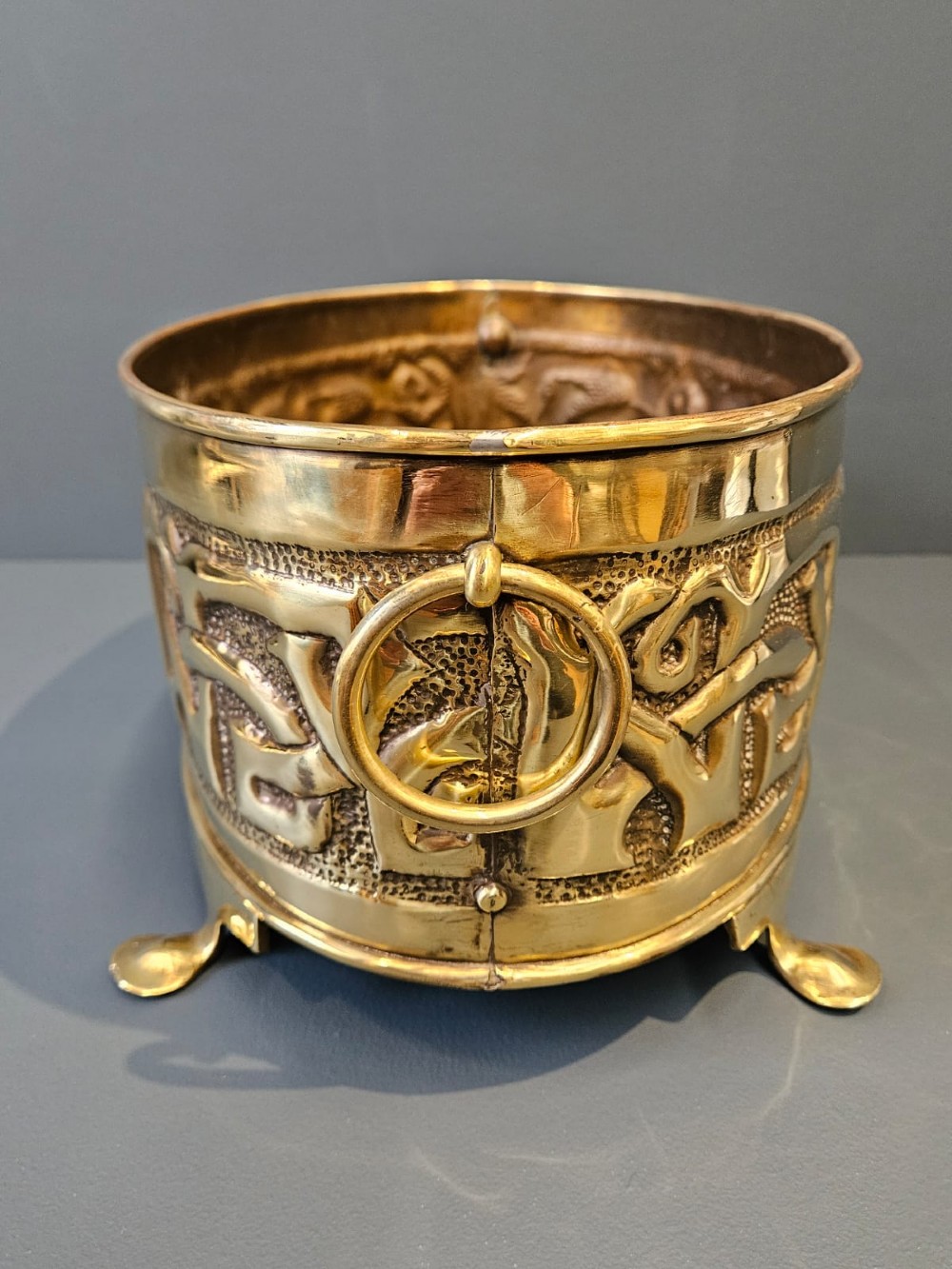 An oval polished brass planter featuring embossed Glasgow School Celtic knotwork on a stippled background, raised on short bracket feet with side handles. View III