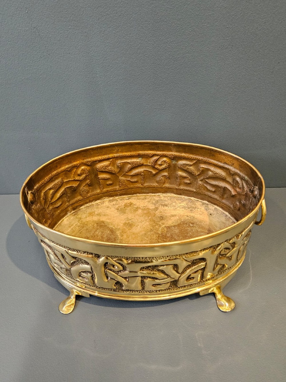An oval polished brass planter featuring embossed Glasgow School Celtic knotwork on a stippled background, raised on short bracket feet with side handles. View V