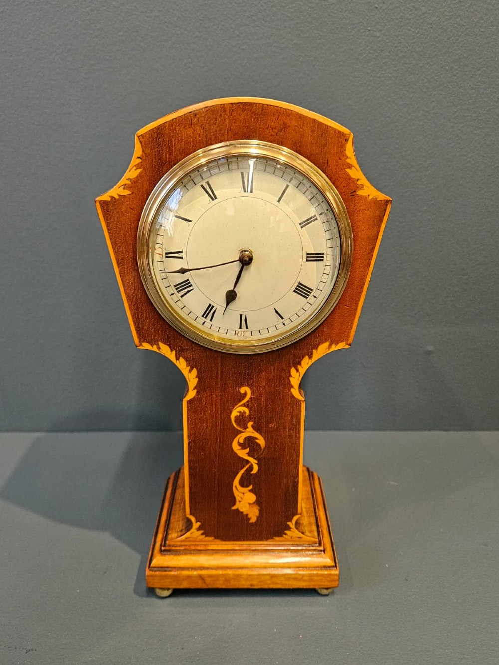 An Edwardian waisted mahogany mantel clock featuring light wood marquetry inlay, a glazed brass bezel, and Roman numerals on a white dial.