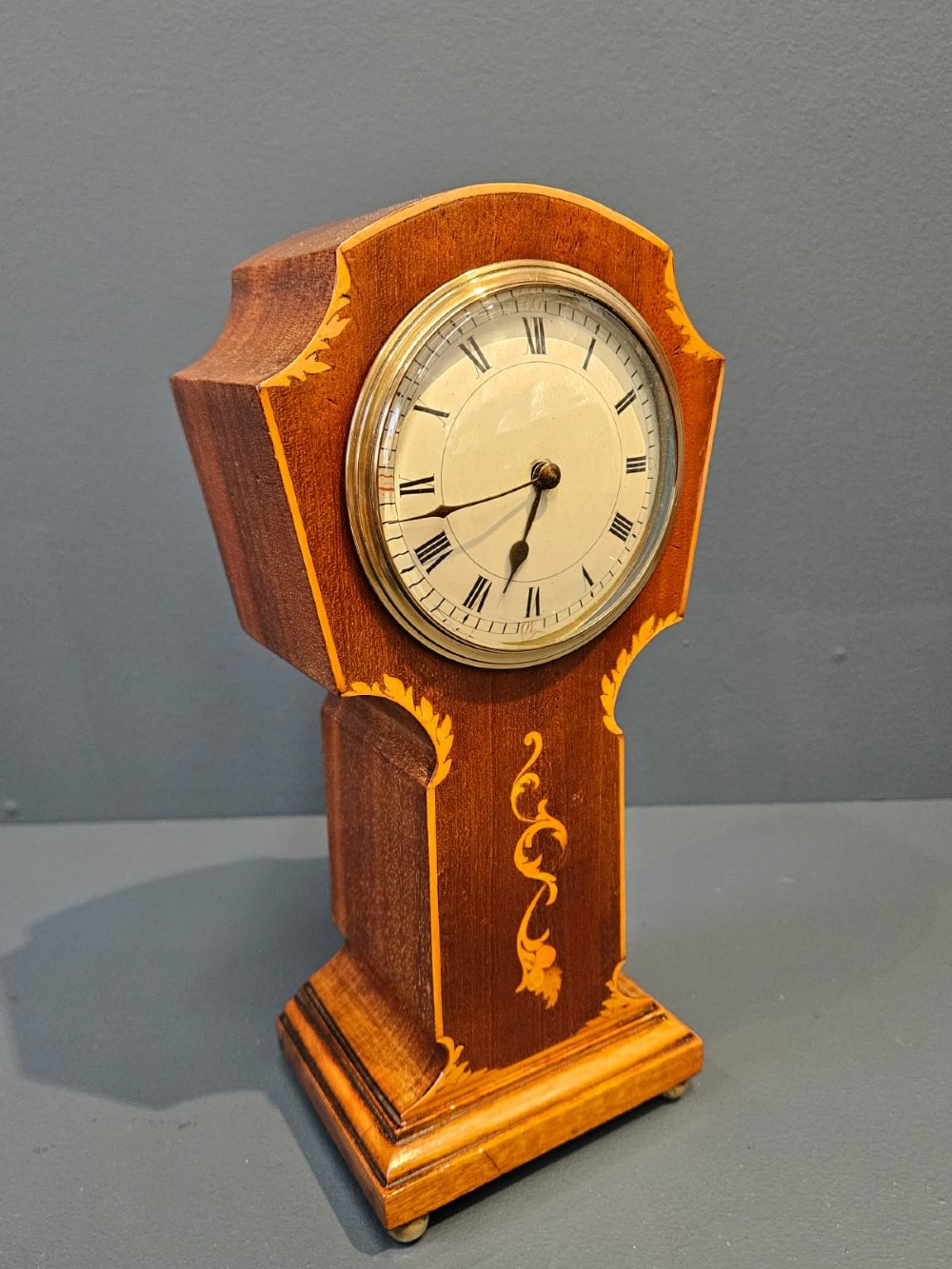 An Edwardian waisted mahogany mantel clock featuring light wood marquetry inlay, a glazed brass bezel, and Roman numerals on a white dial. View II