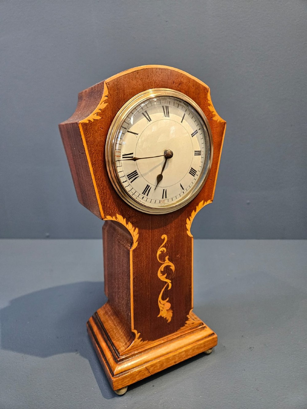 An Edwardian waisted mahogany mantel clock featuring light wood marquetry inlay, a glazed brass bezel, and Roman numerals on a white dial. View V