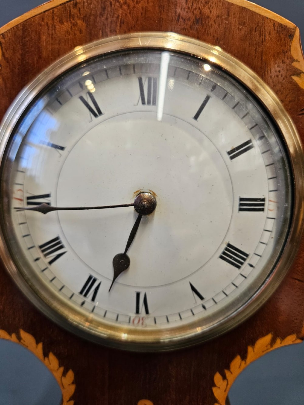 An Edwardian waisted mahogany mantel clock featuring light wood marquetry inlay, a glazed brass bezel, and Roman numerals on a white dial. View VI