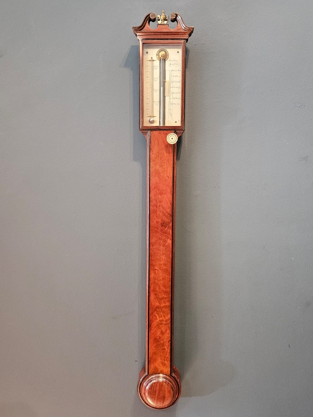 A slim vertical Georgian mahogany stick barometer featuring a broken-arch pediment with a brass finial, an engraved silvered register plate, and a turned cistern cover at the base.