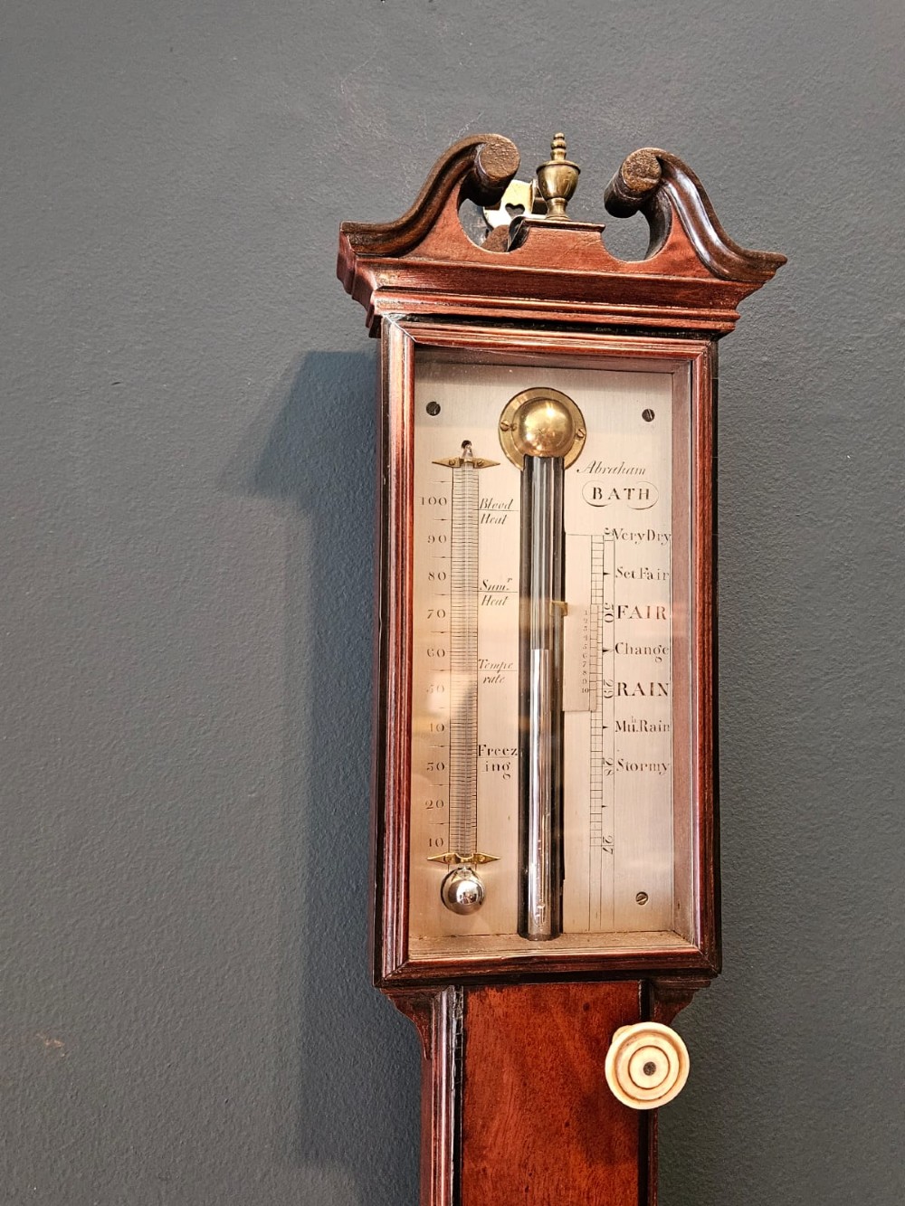A slim vertical Georgian mahogany stick barometer featuring a broken-arch pediment with a brass finial, an engraved silvered register plate, and a turned cistern cover at the base. View III