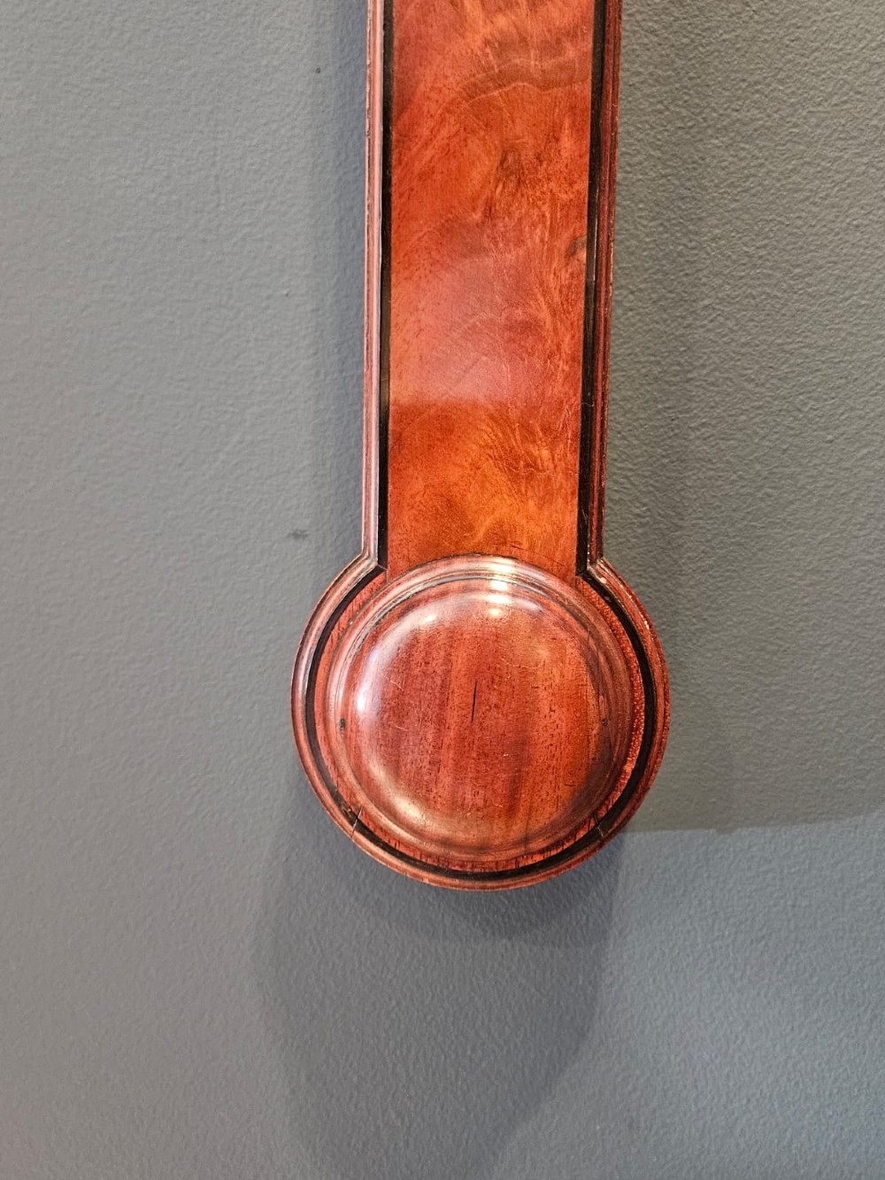 A slim vertical Georgian mahogany stick barometer featuring a broken-arch pediment with a brass finial, an engraved silvered register plate, and a turned cistern cover at the base. View IV