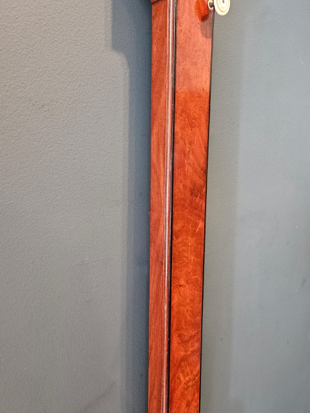 A slim vertical Georgian mahogany stick barometer featuring a broken-arch pediment with a brass finial, an engraved silvered register plate, and a turned cistern cover at the base. View VI