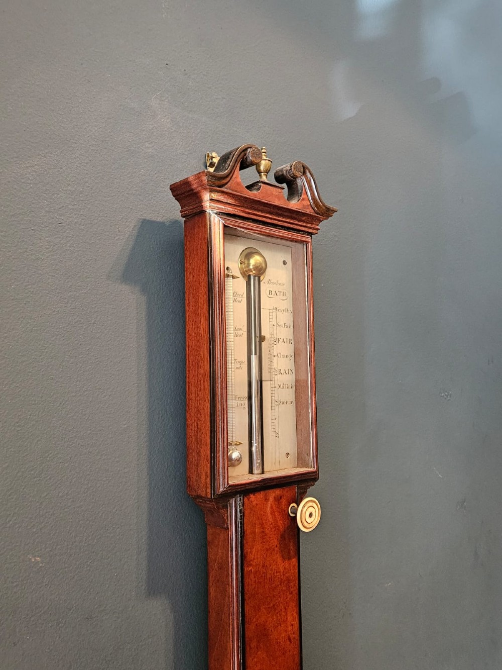 A slim vertical Georgian mahogany stick barometer featuring a broken-arch pediment with a brass finial, an engraved silvered register plate, and a turned cistern cover at the base. View VII
