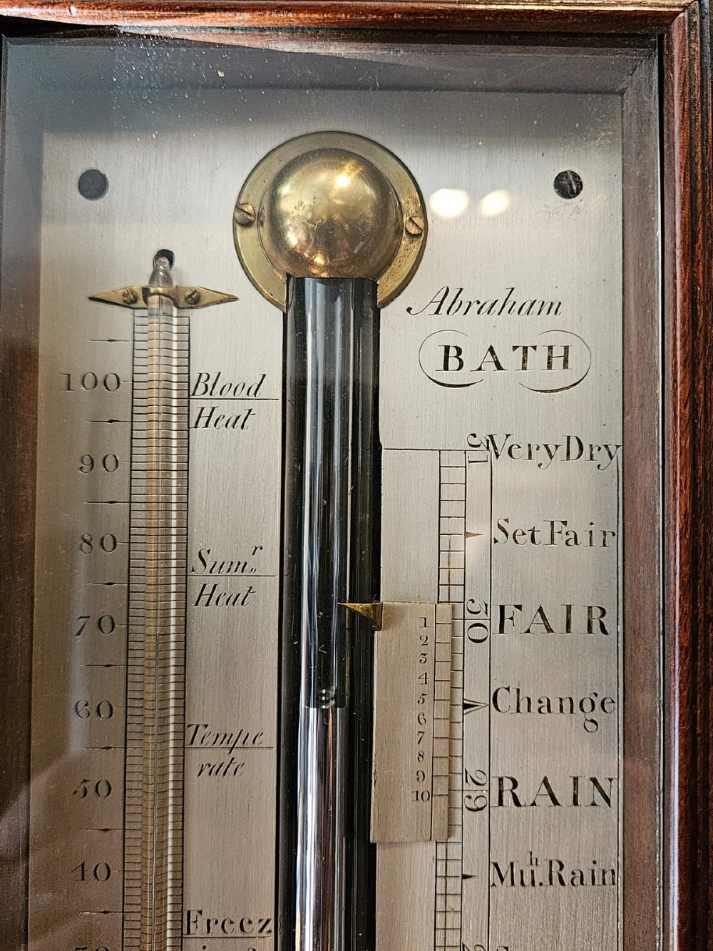 A slim vertical Georgian mahogany stick barometer featuring a broken-arch pediment with a brass finial, an engraved silvered register plate, and a turned cistern cover at the base. View VIII