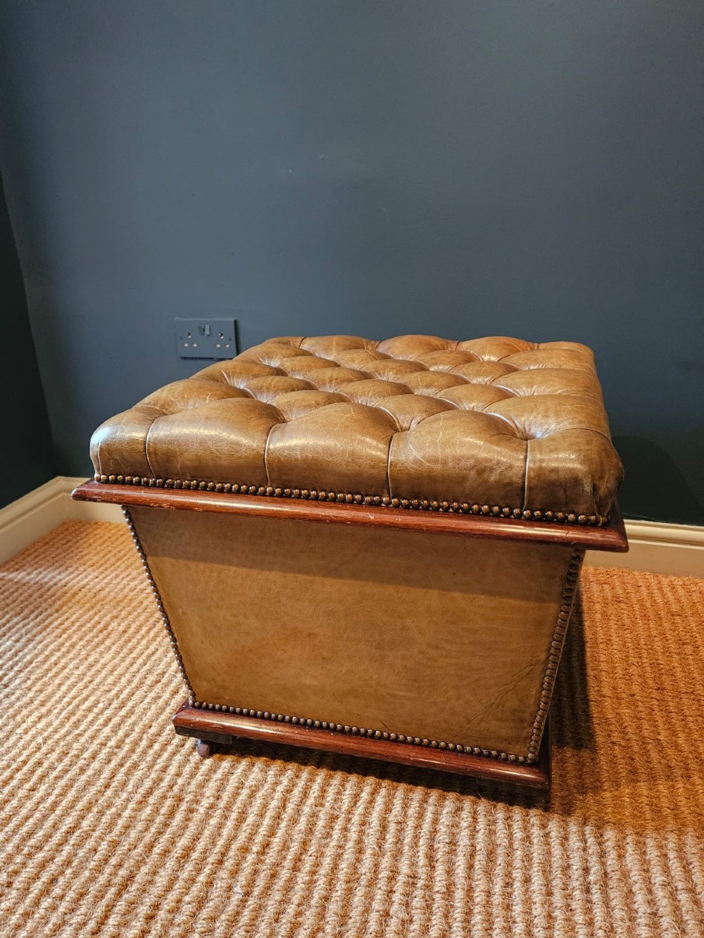 A deep-buttoned tan leather Victorian ottoman stool featuring brass stud trim and set upon a mahogany base with castors.
