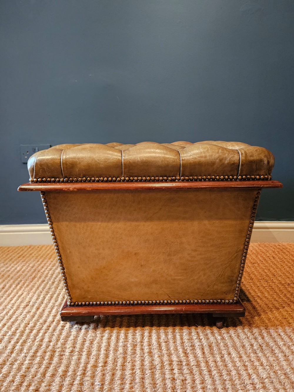 A deep-buttoned tan leather Victorian ottoman stool featuring brass stud trim and set upon a mahogany base with castors. View VI