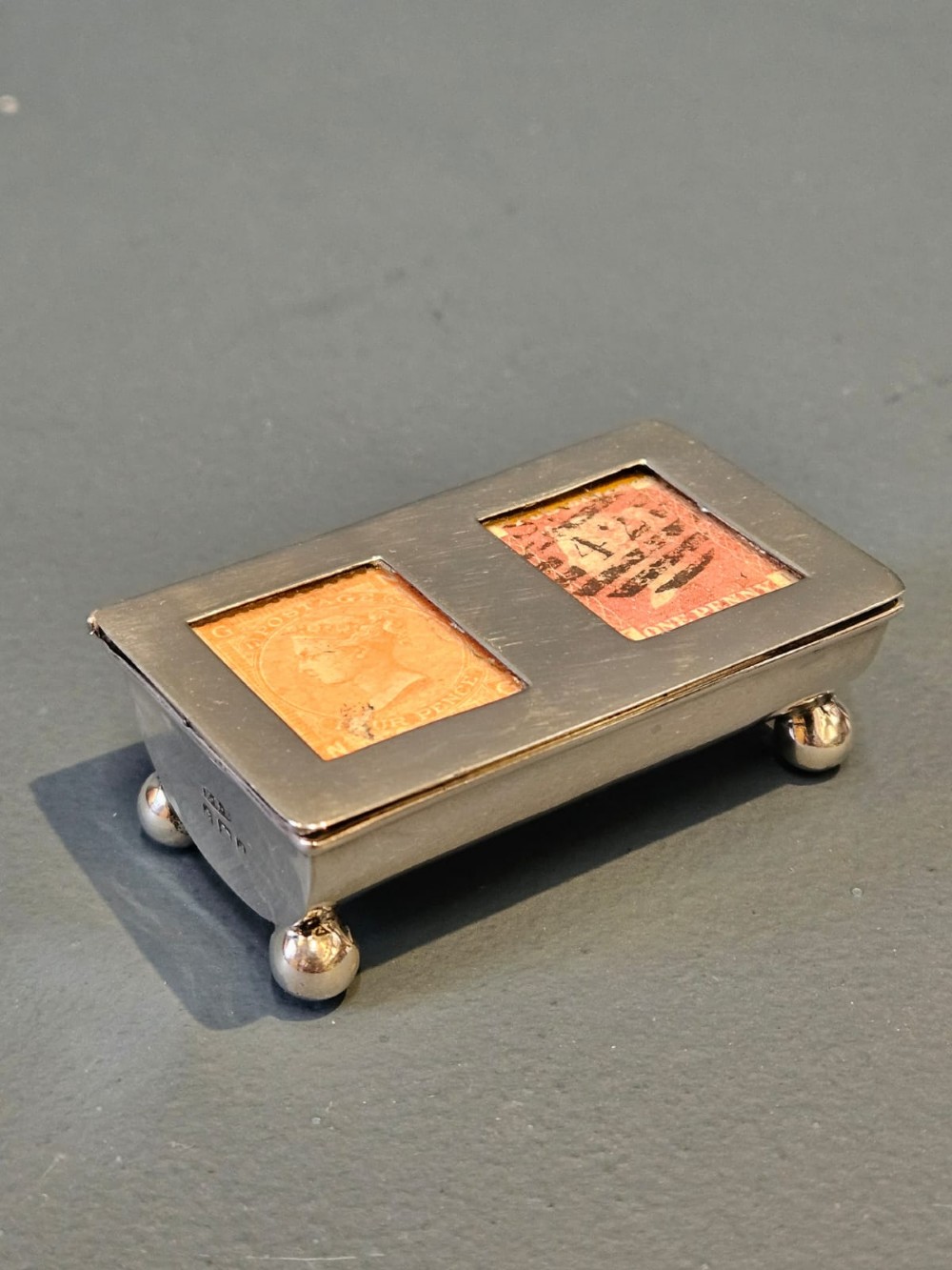 A small rectangular Victorian silver stamp box raised on four ball feet with two glass apertures on the lid displaying postage stamps.
