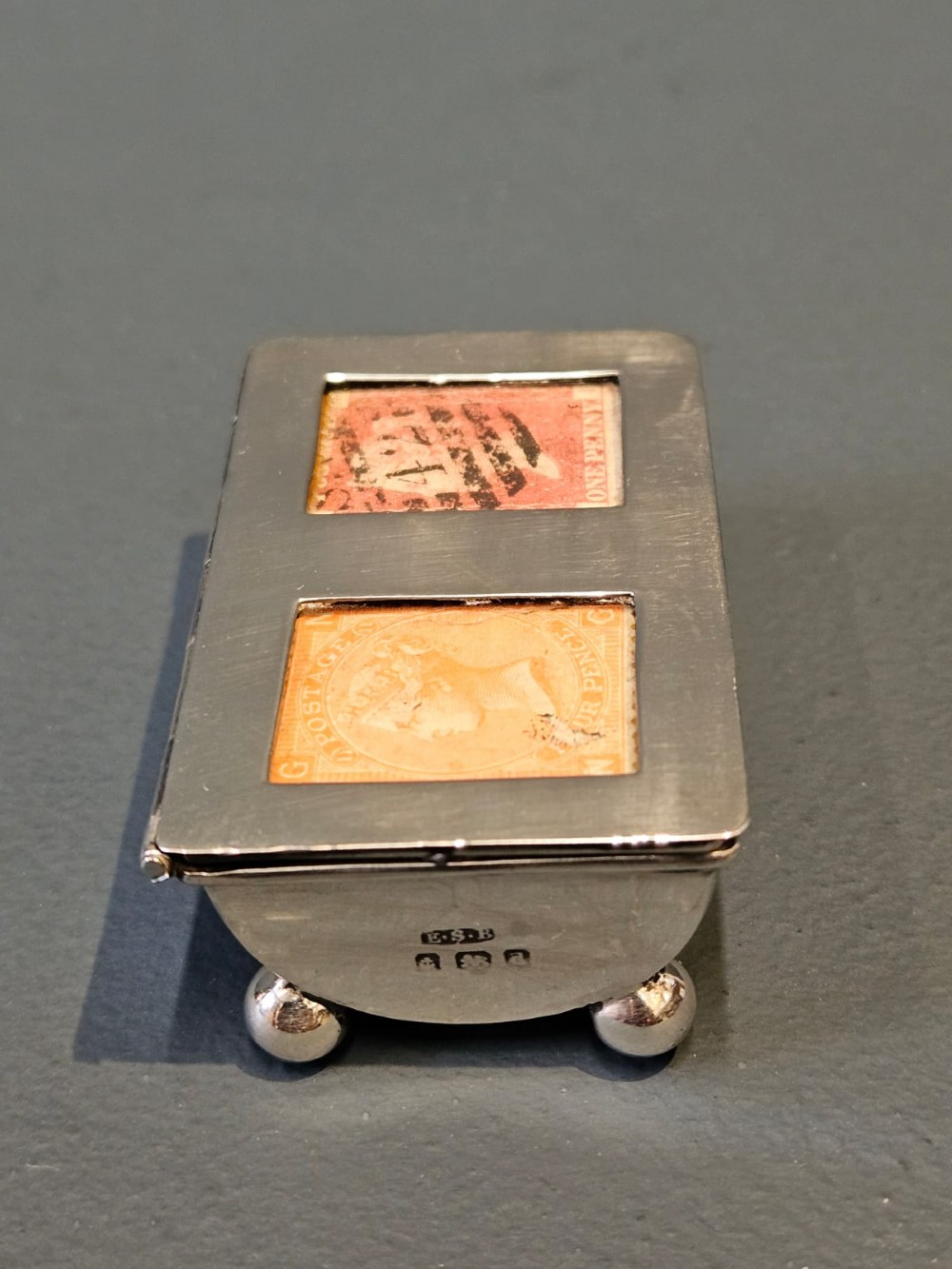 A small rectangular Victorian silver stamp box raised on four ball feet with two glass apertures on the lid displaying postage stamps. View II