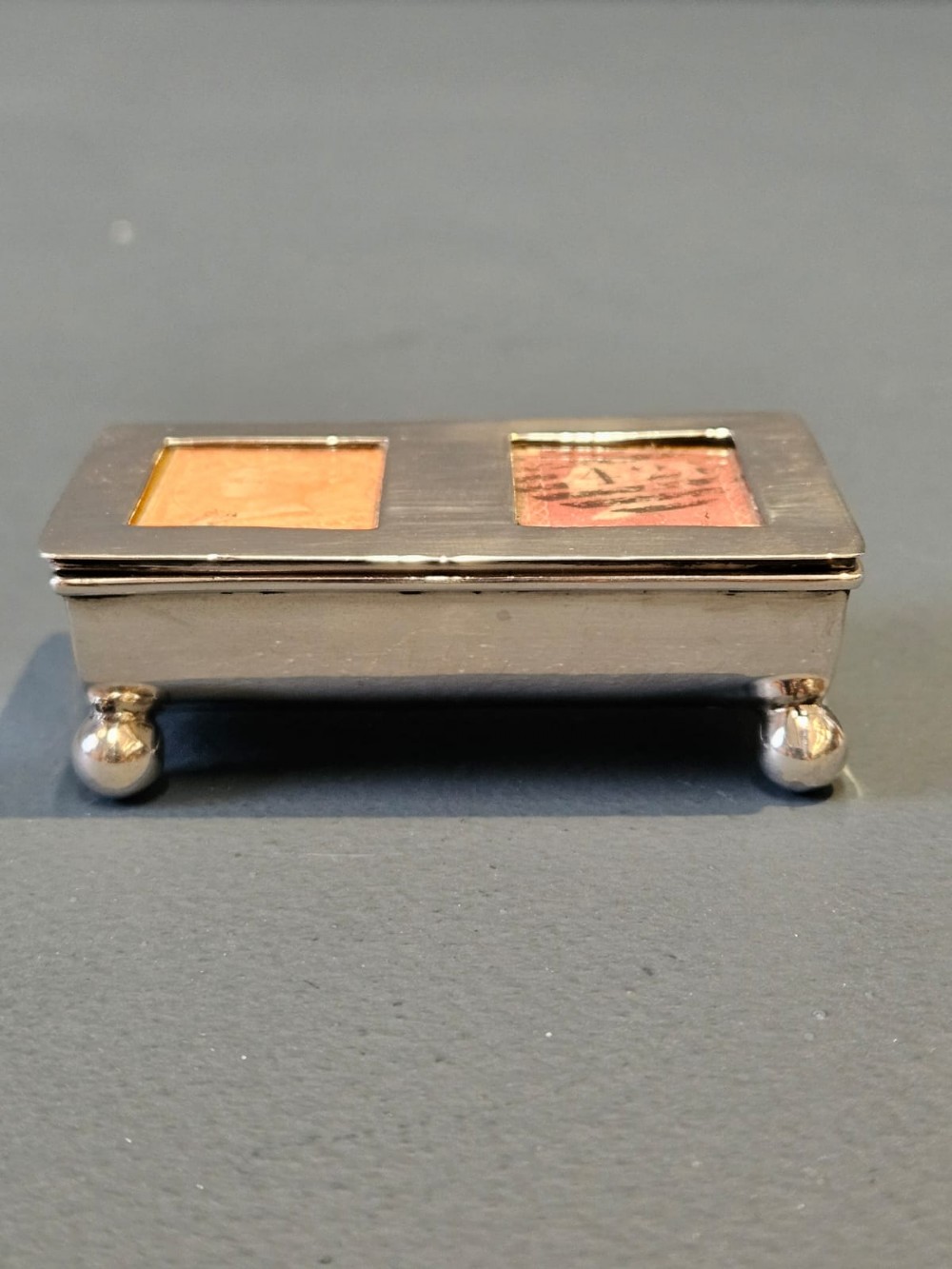 A small rectangular Victorian silver stamp box raised on four ball feet with two glass apertures on the lid displaying postage stamps. View IV