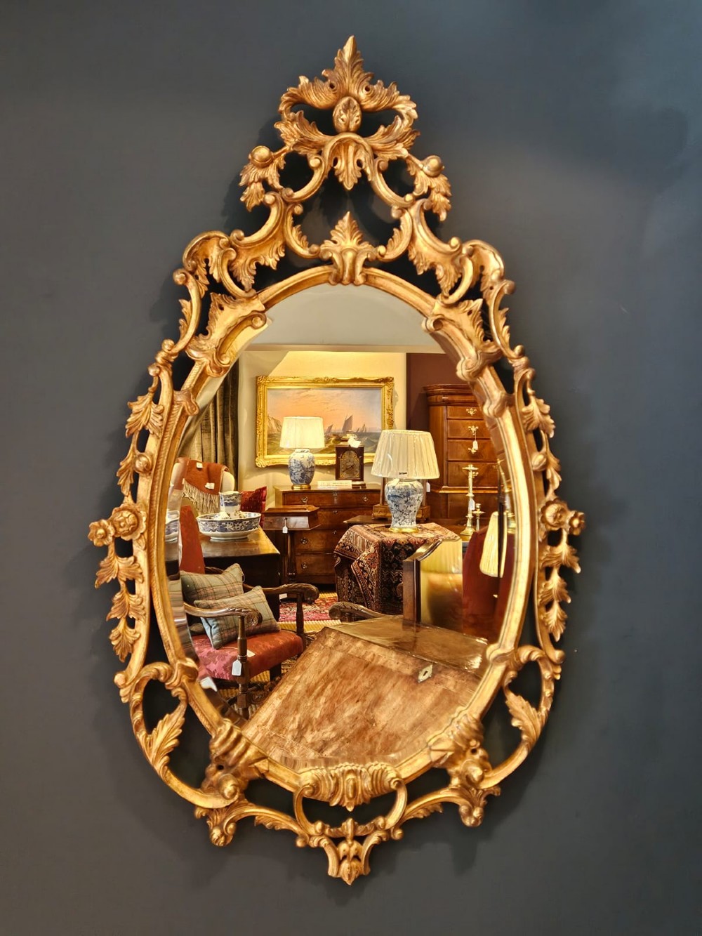 An oval Rococo style wall mirror featuring an elaborate gilded frame decorated with carved acanthus leaf scrollwork and a prominent cresting top.