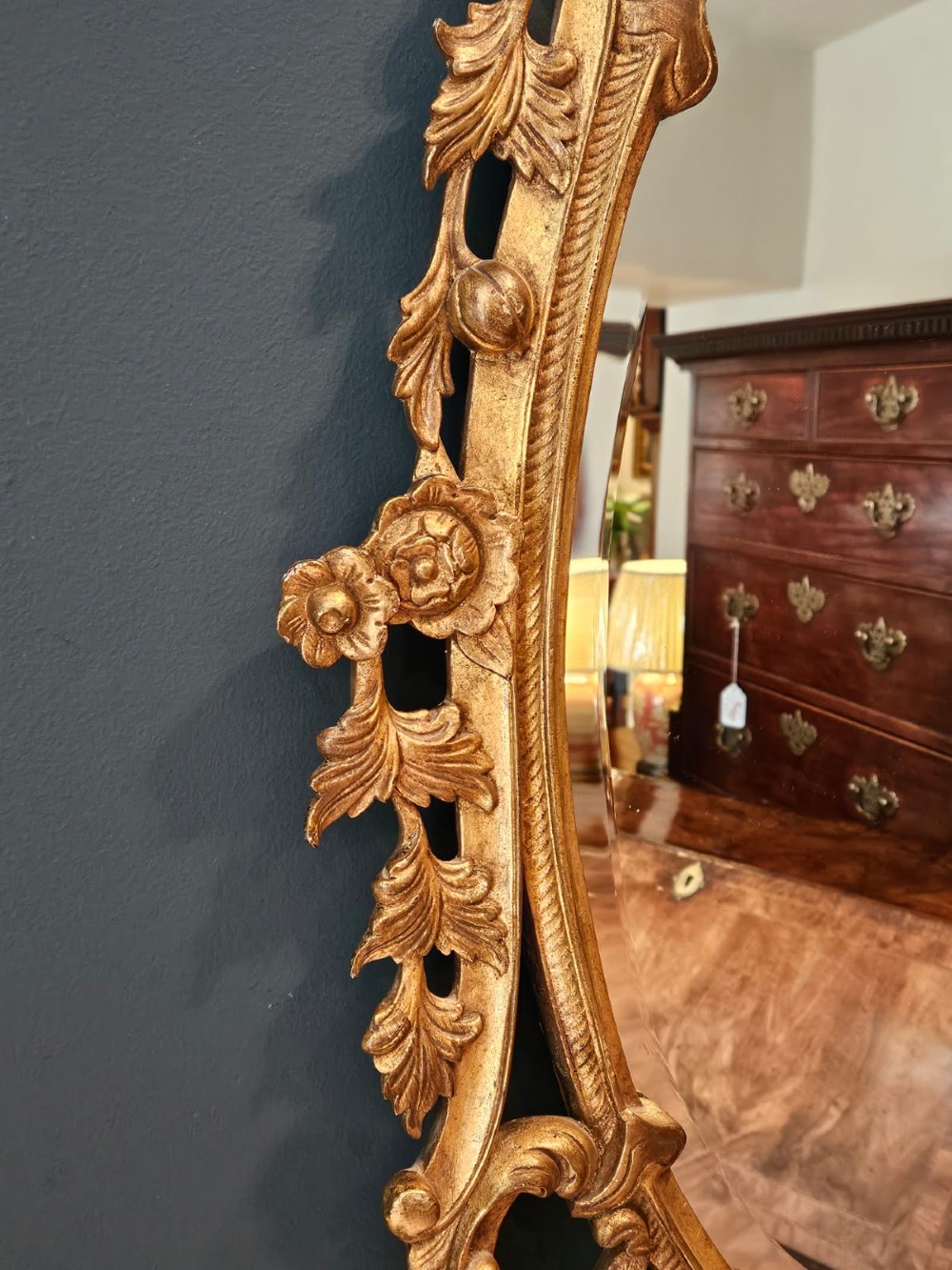 An oval Rococo style wall mirror featuring an elaborate gilded frame decorated with carved acanthus leaf scrollwork and a prominent cresting top. View II