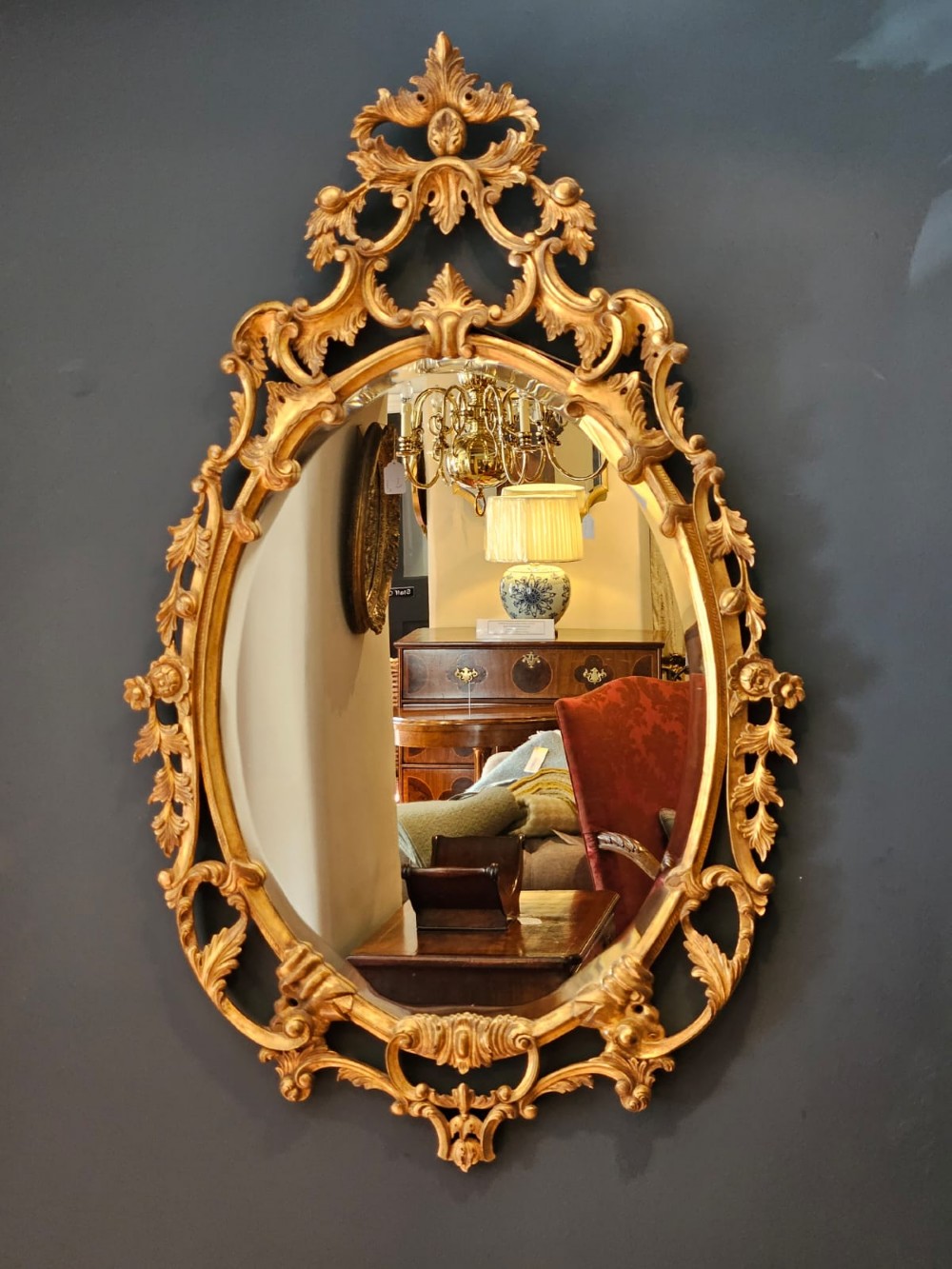 An oval Rococo style wall mirror featuring an elaborate gilded frame decorated with carved acanthus leaf scrollwork and a prominent cresting top. View III