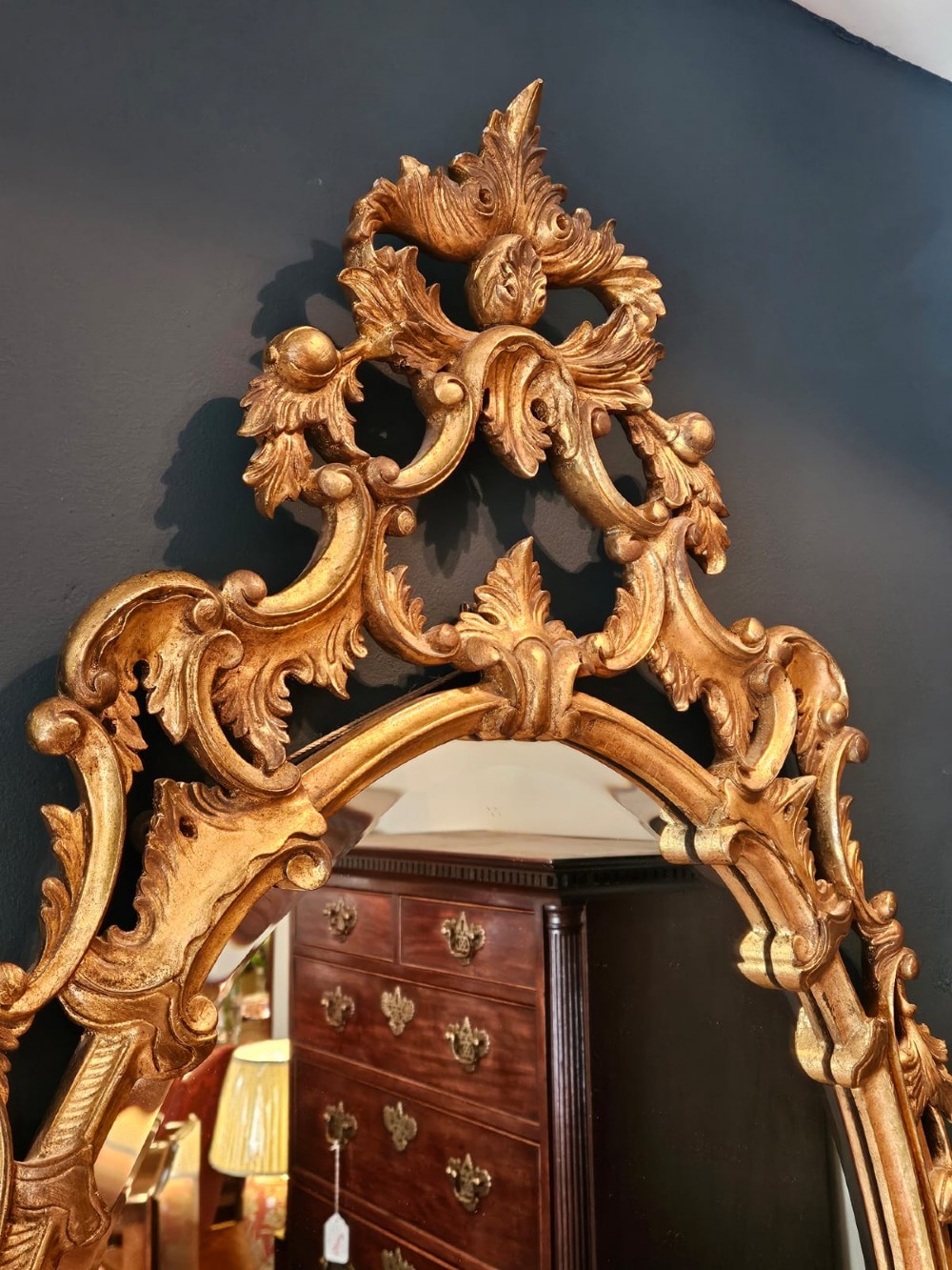 An oval Rococo style wall mirror featuring an elaborate gilded frame decorated with carved acanthus leaf scrollwork and a prominent cresting top. View IV