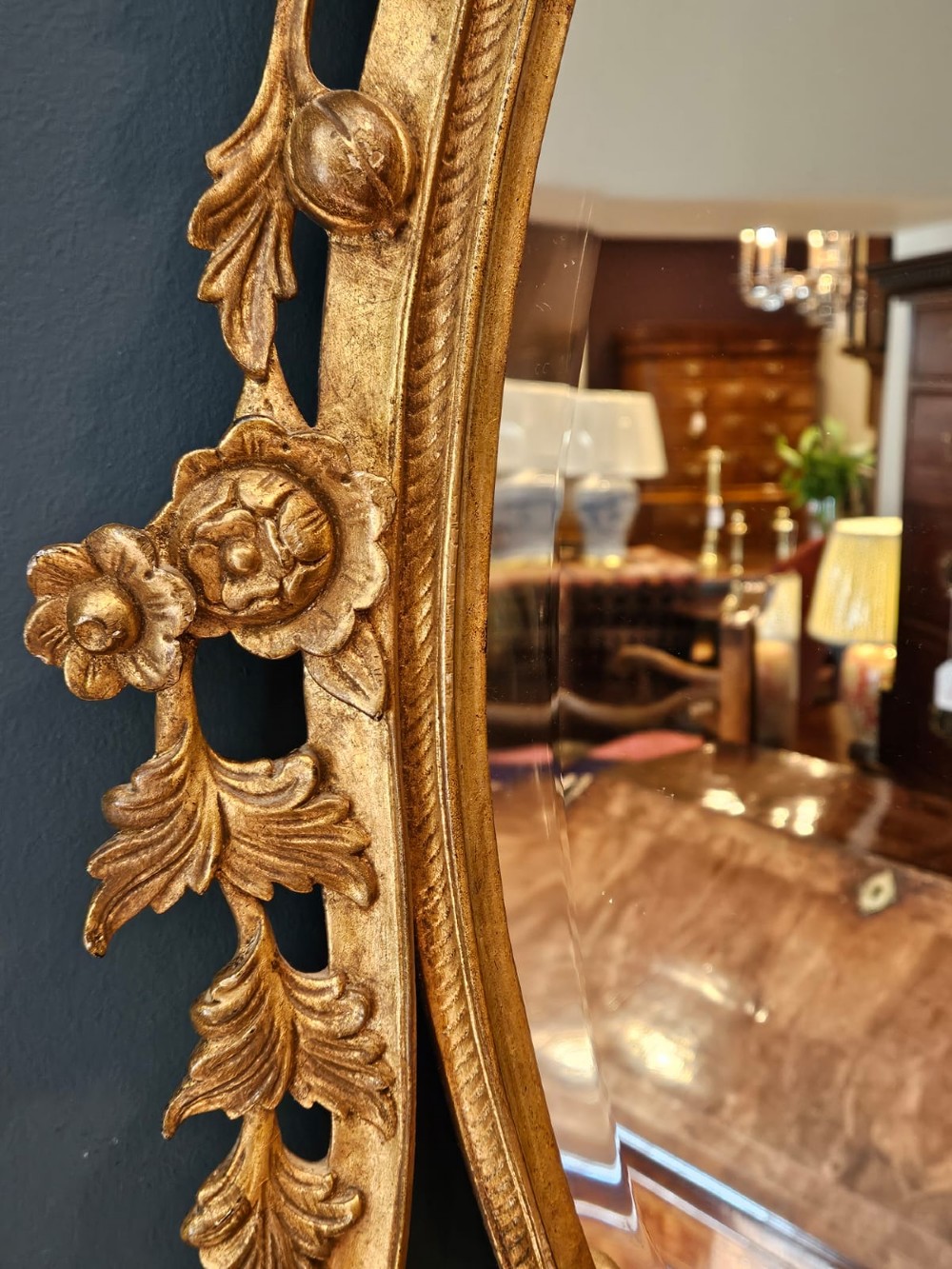 An oval Rococo style wall mirror featuring an elaborate gilded frame decorated with carved acanthus leaf scrollwork and a prominent cresting top. View V