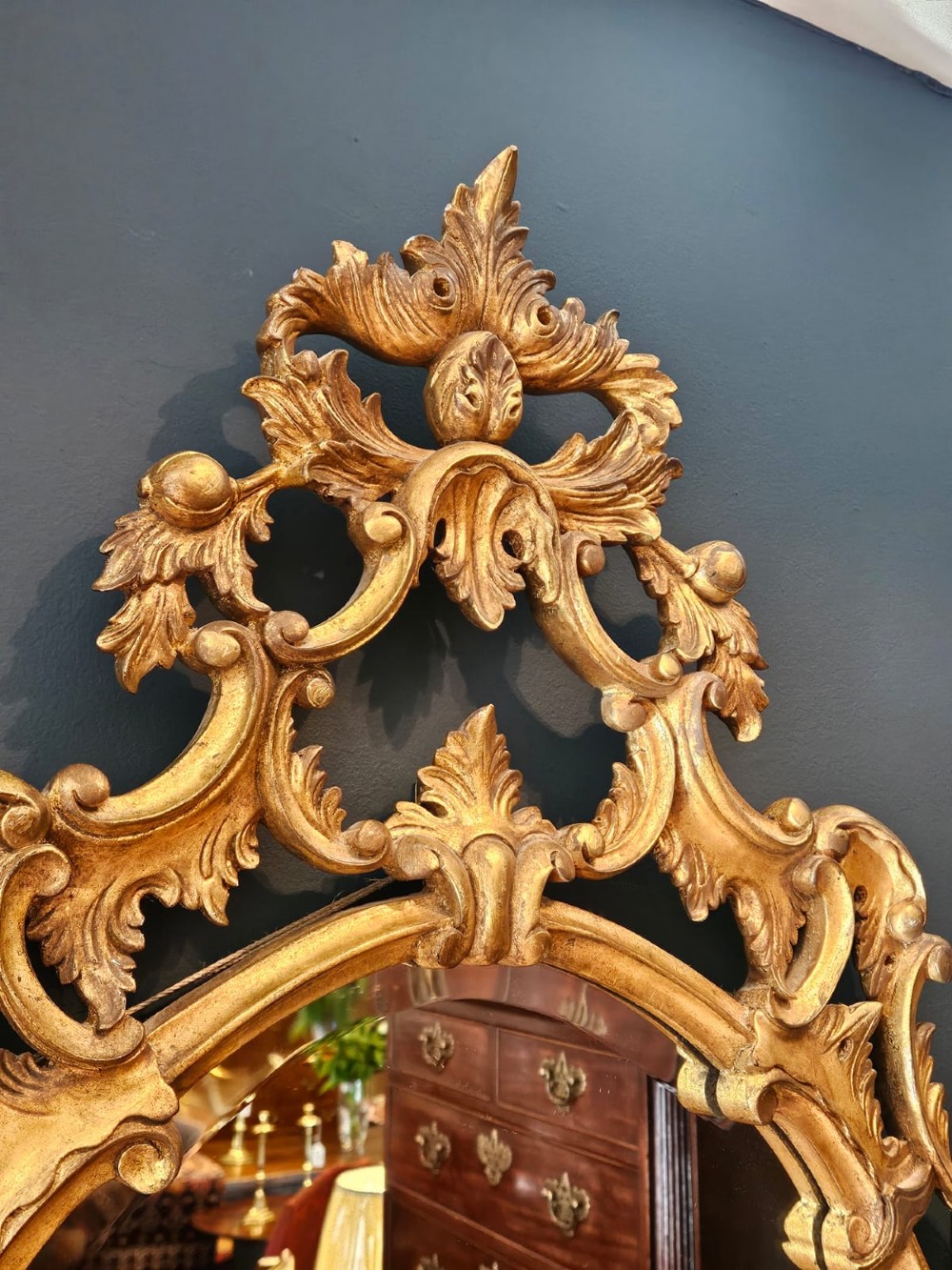 An oval Rococo style wall mirror featuring an elaborate gilded frame decorated with carved acanthus leaf scrollwork and a prominent cresting top. View VI