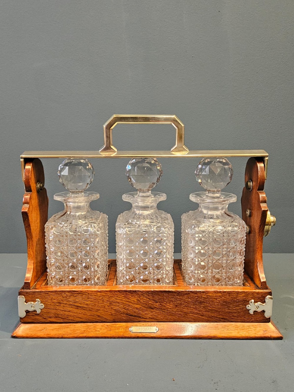 A Victorian oak three-bottle tantalus featuring a wooden frame with silver-plated mounts and three cut-glass square decanters with round faceted stoppers.