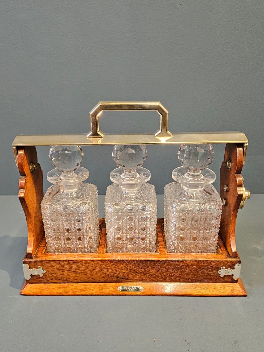 A Victorian oak three-bottle tantalus featuring a wooden frame with silver-plated mounts and three cut-glass square decanters with round faceted stoppers. View II