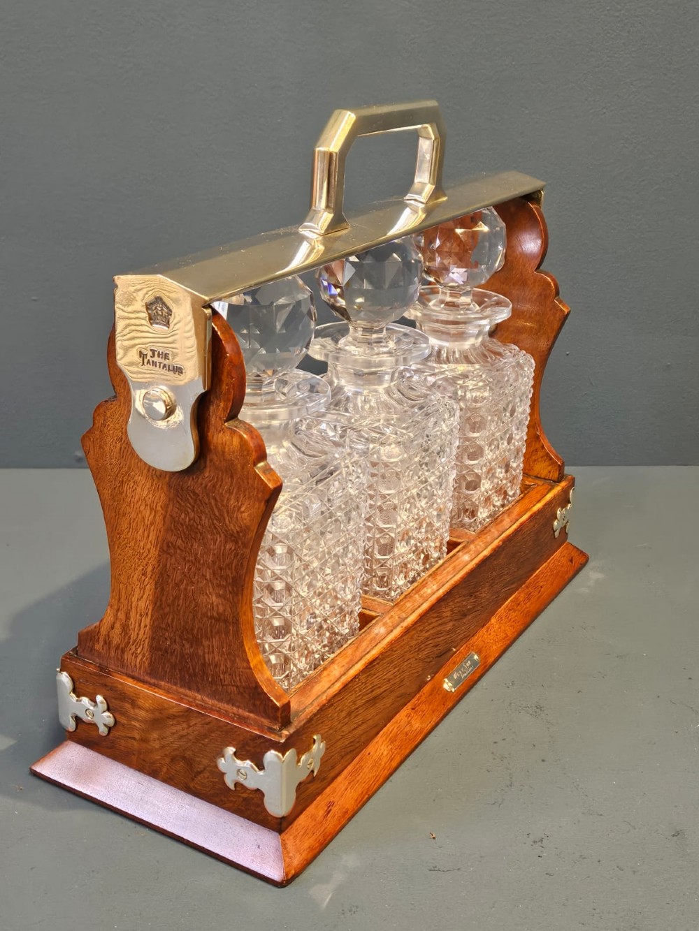 A Victorian oak three-bottle tantalus featuring a wooden frame with silver-plated mounts and three cut-glass square decanters with round faceted stoppers. View V