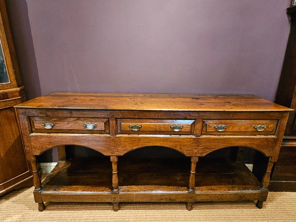 A George III oak open pot board dresser base featuring three drawers with brass drop handles, a shaped arched apron, and turned legs.