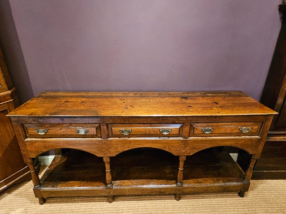 A George III oak open pot board dresser base featuring three drawers with brass drop handles, a shaped arched apron, and turned legs. View II