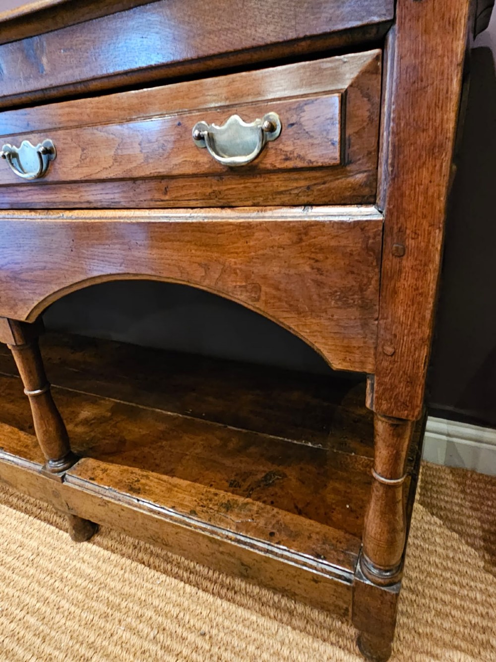 A George III oak open pot board dresser base featuring three drawers with brass drop handles, a shaped arched apron, and turned legs. View V