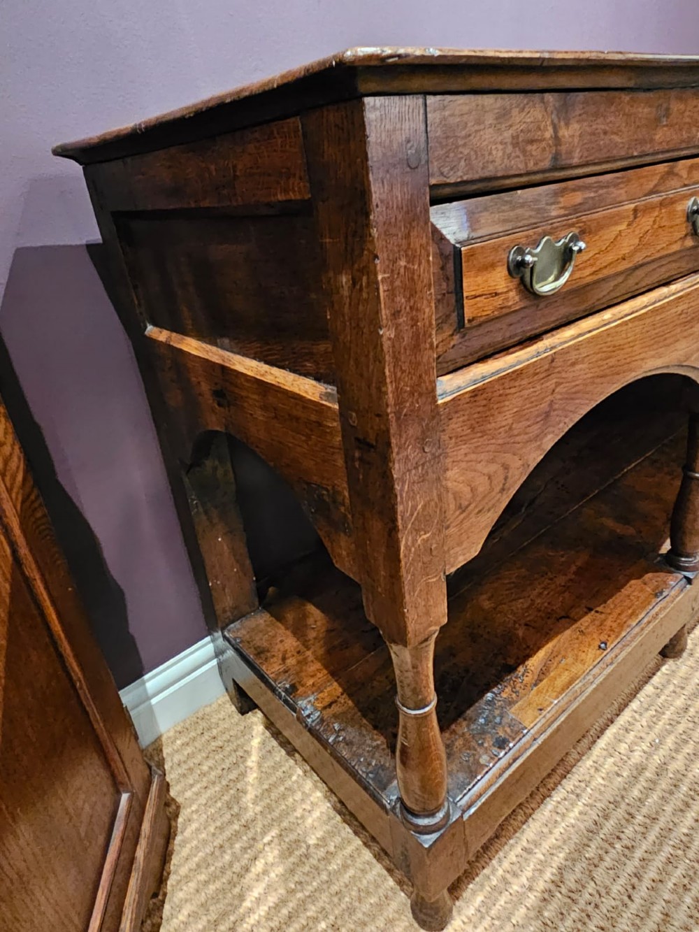 A George III oak open pot board dresser base featuring three drawers with brass drop handles, a shaped arched apron, and turned legs. View X