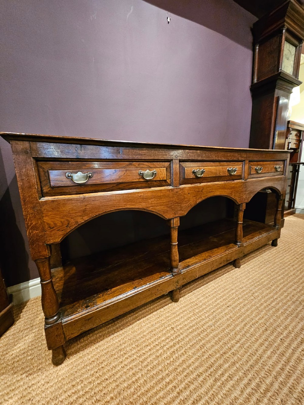 A George III oak open pot board dresser base featuring three drawers with brass drop handles, a shaped arched apron, and turned legs. View XI