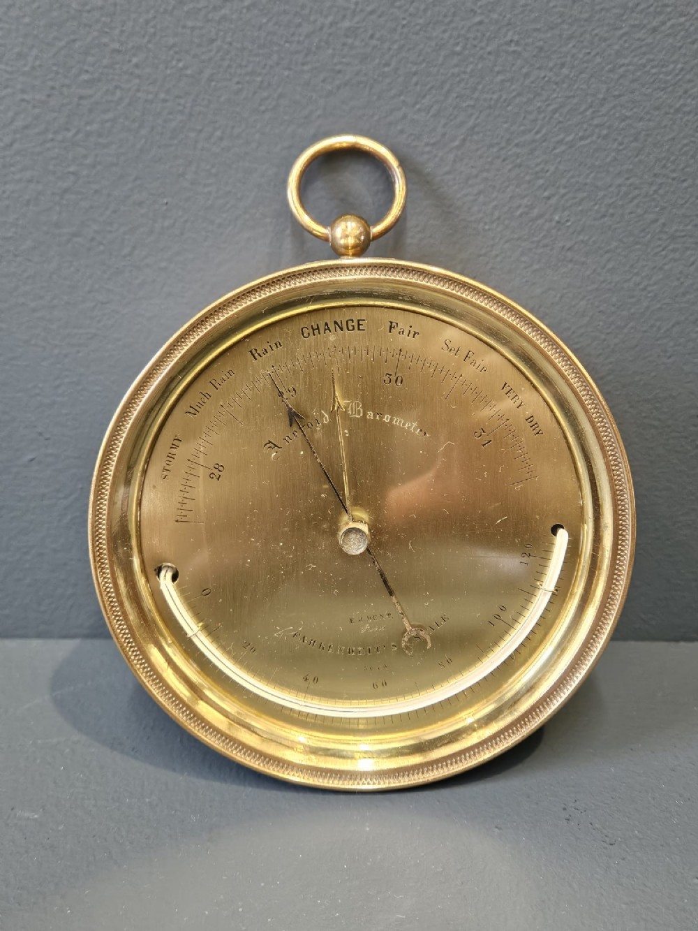 A circular Victorian brass aneroid barometer featuring an engraved face with weather markings, an integrated thermometer, and a top hanging ring.