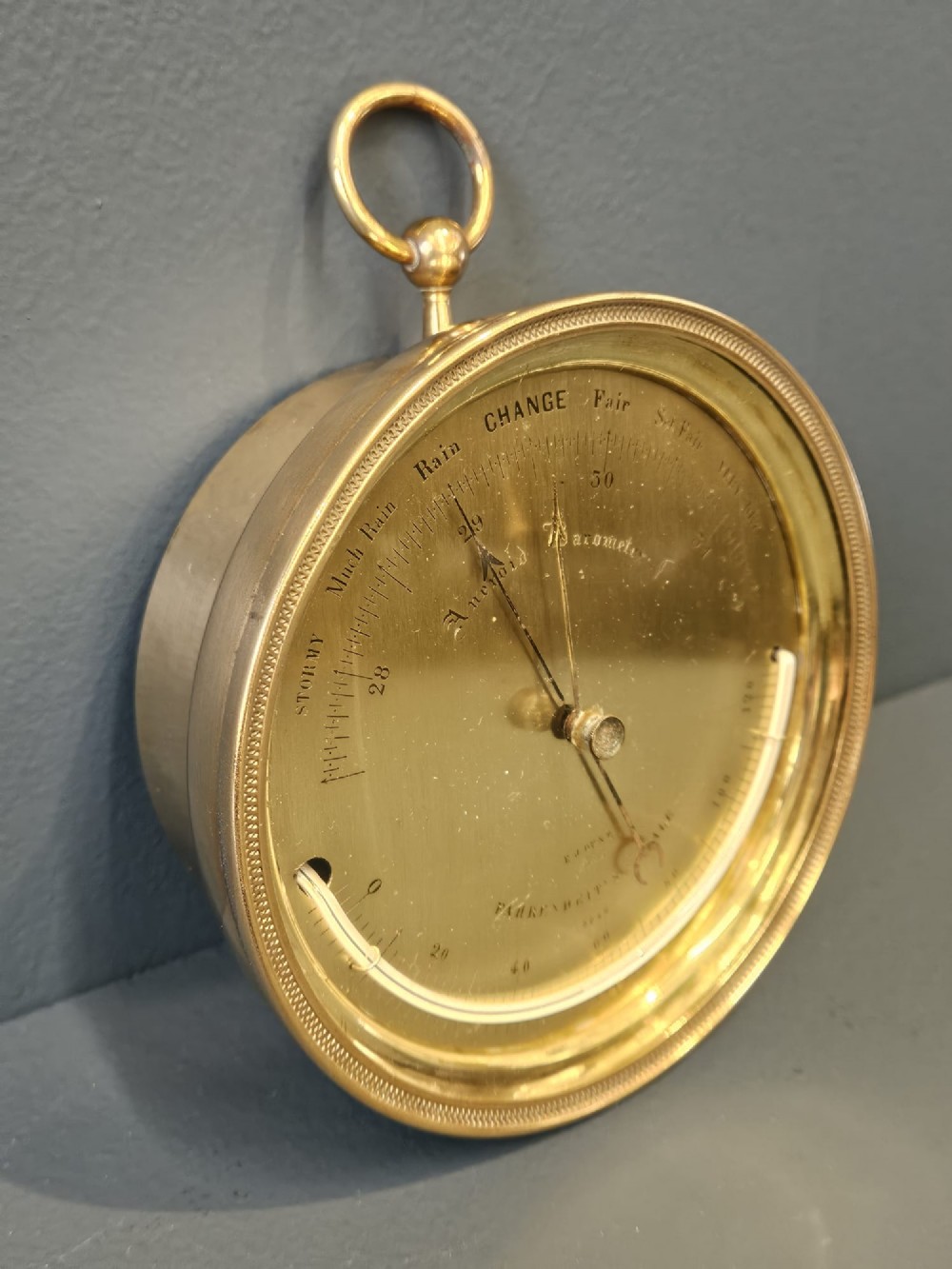 A circular Victorian brass aneroid barometer featuring an engraved face with weather markings, an integrated thermometer, and a top hanging ring. View III