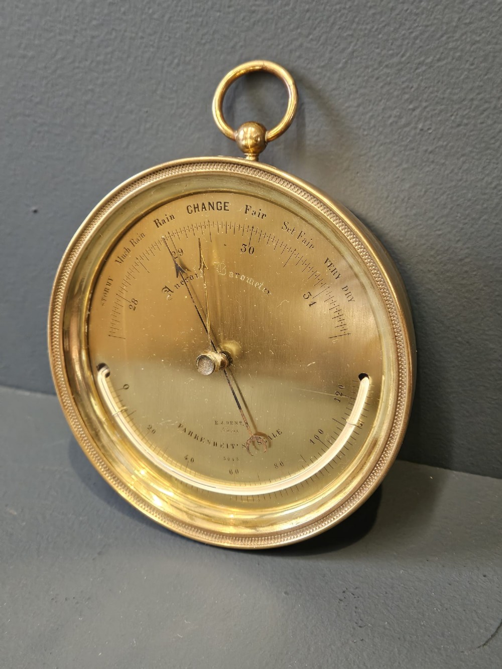 A circular Victorian brass aneroid barometer featuring an engraved face with weather markings, an integrated thermometer, and a top hanging ring. View V