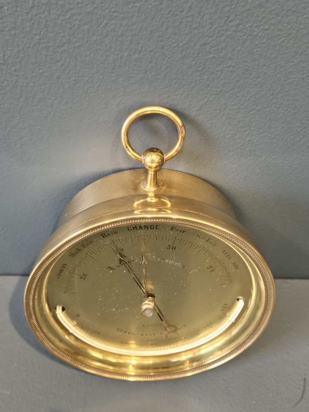 A circular Victorian brass aneroid barometer featuring an engraved face with weather markings, an integrated thermometer, and a top hanging ring. View VI