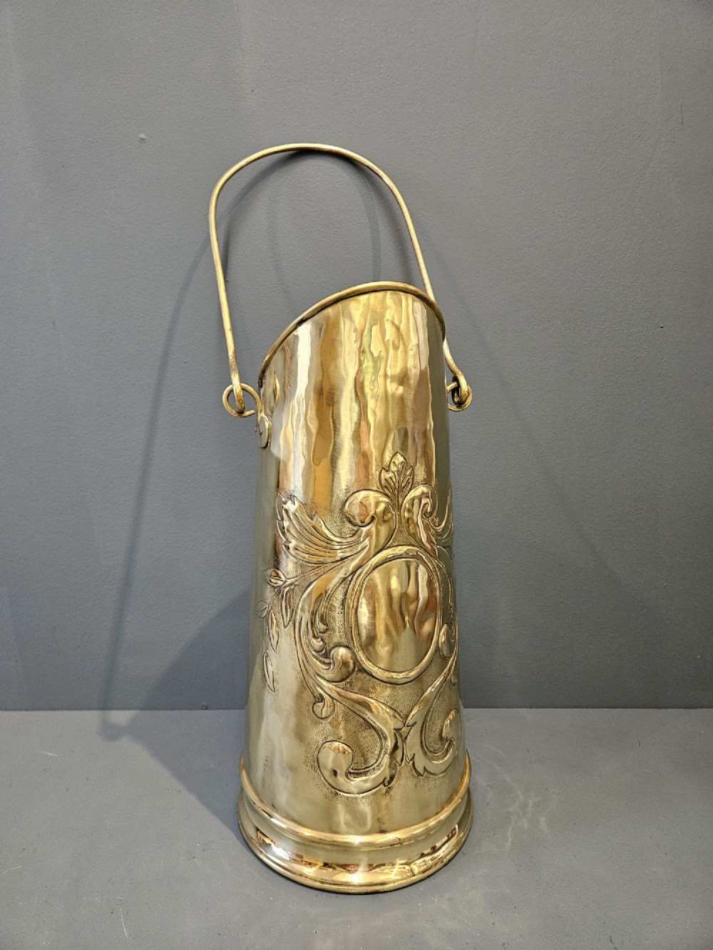 A tall, polished brass cylindrical coal scuttle featuring an arched swing handle and embossed Art Nouveau scroll motifs on the front.