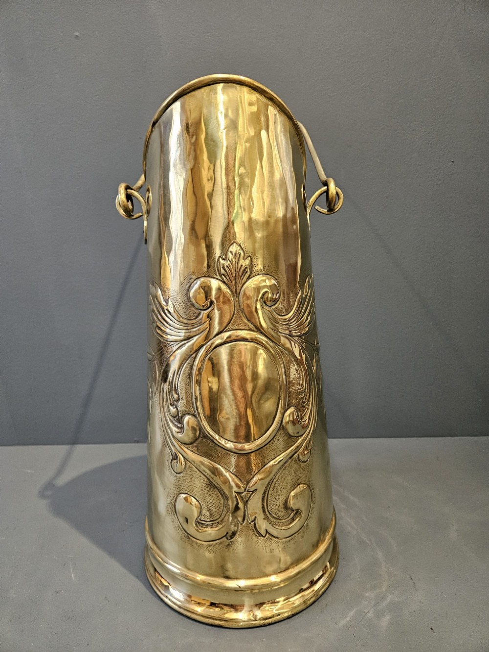 A tall, polished brass cylindrical coal scuttle featuring an arched swing handle and embossed Art Nouveau scroll motifs on the front. View II