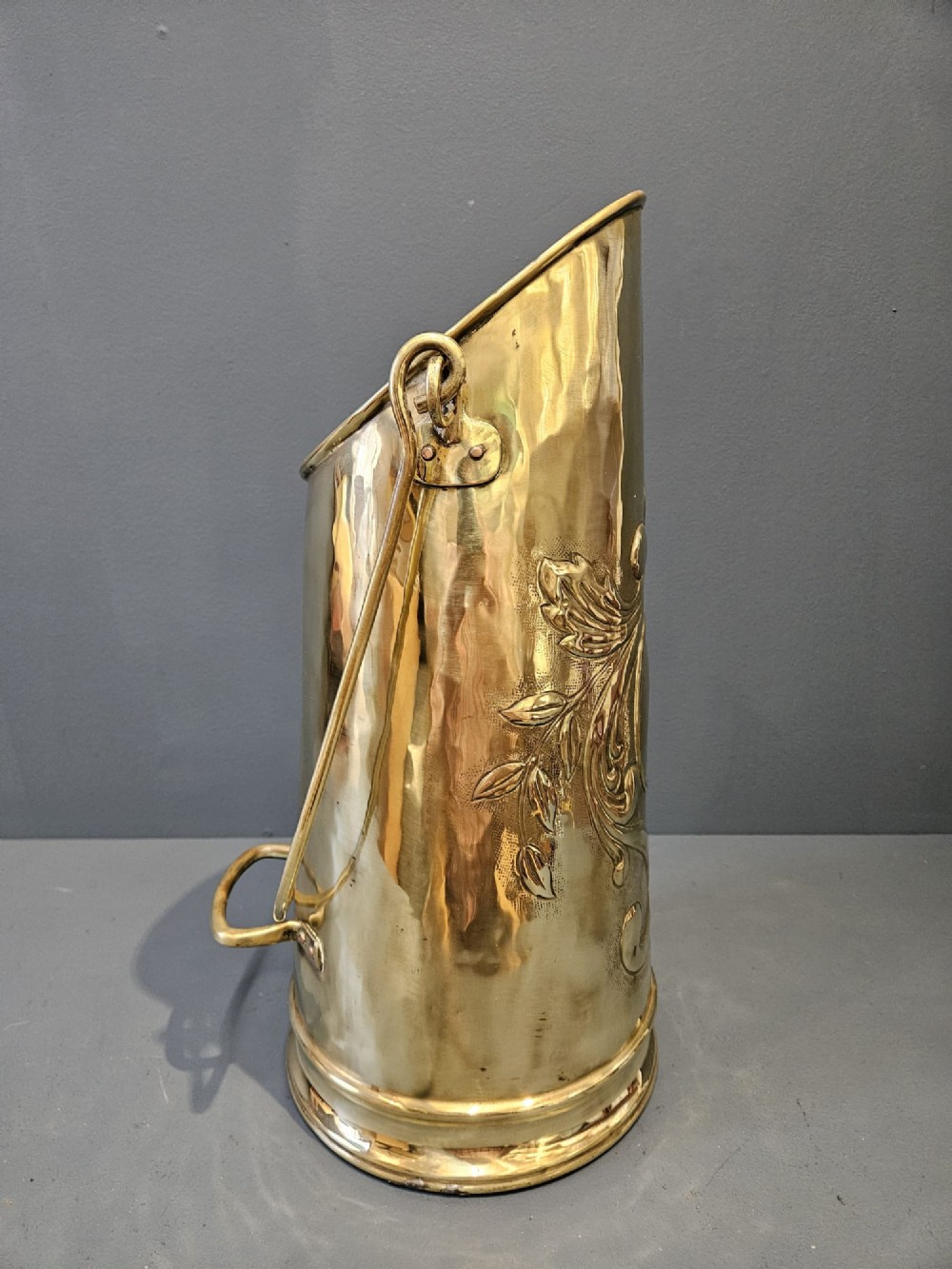 A tall, polished brass cylindrical coal scuttle featuring an arched swing handle and embossed Art Nouveau scroll motifs on the front. View III