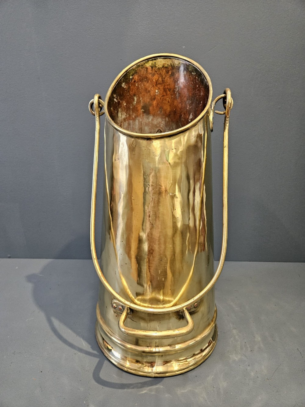 A tall, polished brass cylindrical coal scuttle featuring an arched swing handle and embossed Art Nouveau scroll motifs on the front. View IV