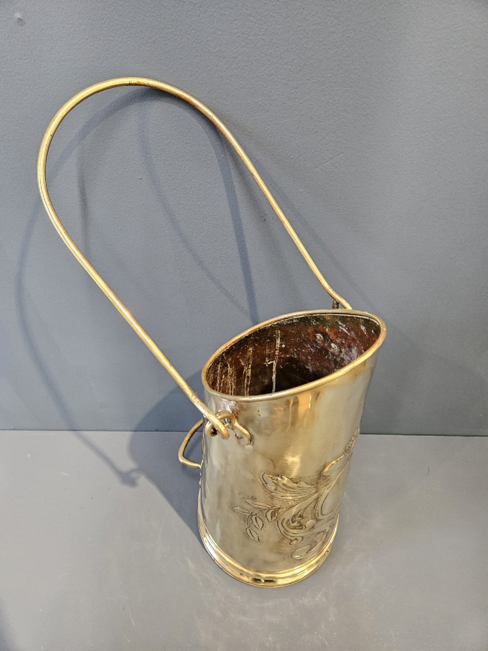 A tall, polished brass cylindrical coal scuttle featuring an arched swing handle and embossed Art Nouveau scroll motifs on the front. View V