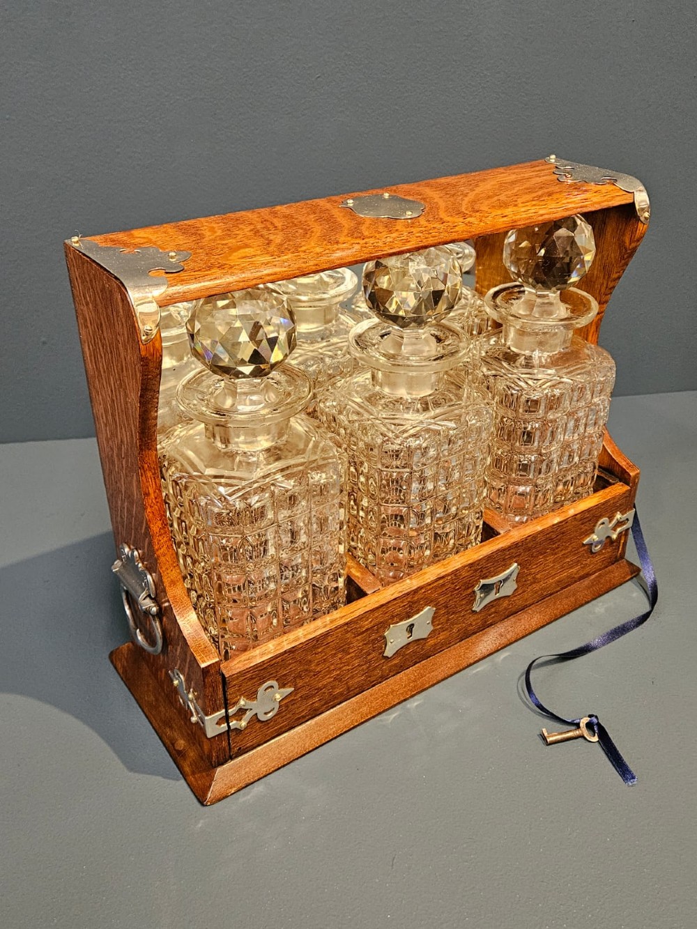 A Victorian oak three-bottle tantalus featuring silver-plated corner mounts, side handles, front keyholes, and three cut-glass decanters with multi-faceted stoppers.