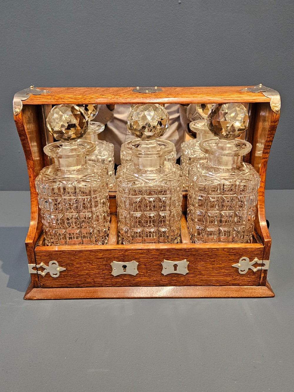 A Victorian oak three-bottle tantalus featuring silver-plated corner mounts, side handles, front keyholes, and three cut-glass decanters with multi-faceted stoppers. View II