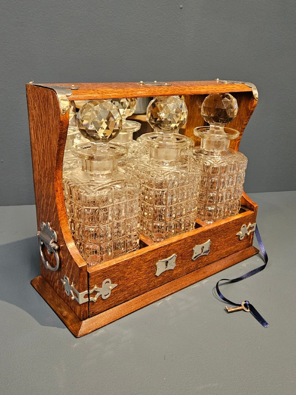 A Victorian oak three-bottle tantalus featuring silver-plated corner mounts, side handles, front keyholes, and three cut-glass decanters with multi-faceted stoppers. View V