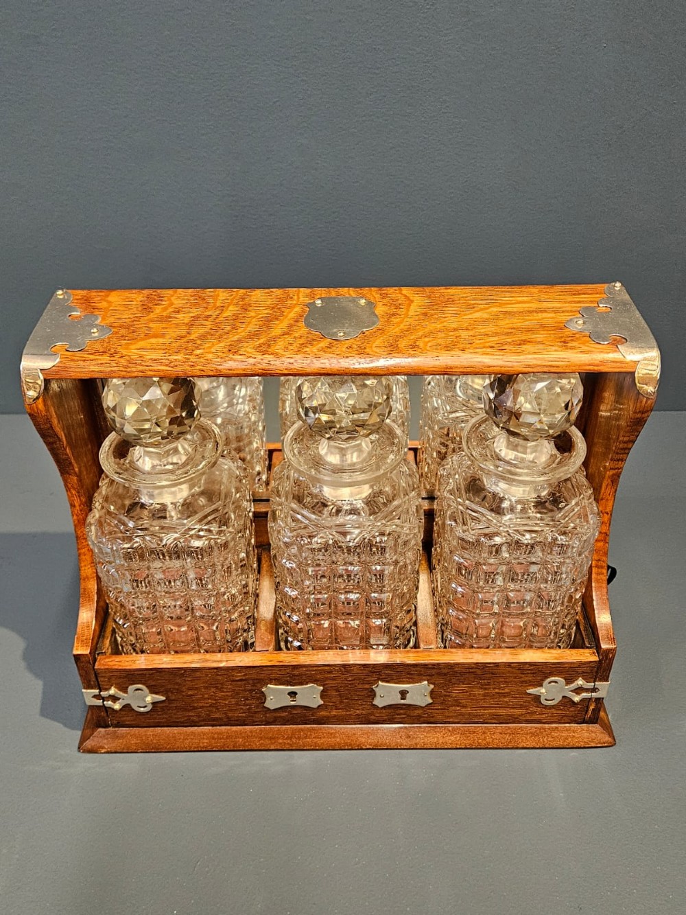 A Victorian oak three-bottle tantalus featuring silver-plated corner mounts, side handles, front keyholes, and three cut-glass decanters with multi-faceted stoppers. View VI