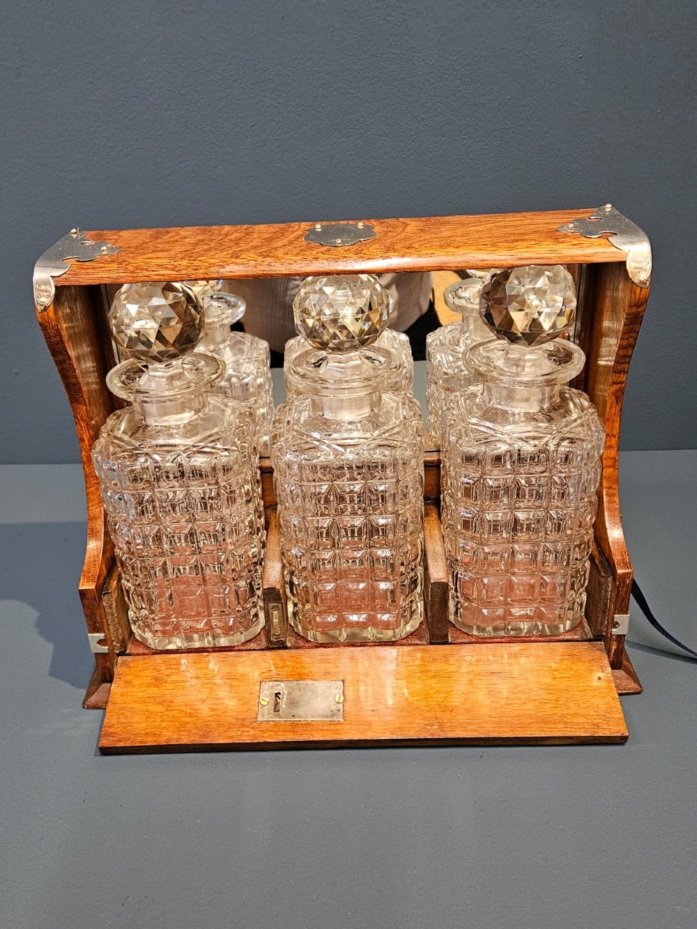 A Victorian oak three-bottle tantalus featuring silver-plated corner mounts, side handles, front keyholes, and three cut-glass decanters with multi-faceted stoppers. View VIII