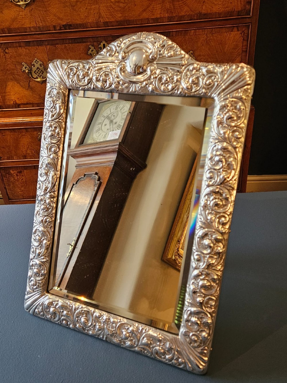 A Victorian easel-back mirror featuring an embossed silver frame decorated with scrollwork, acanthus leaves, and a central cresting cartouche around a bevelled glass plate. View V