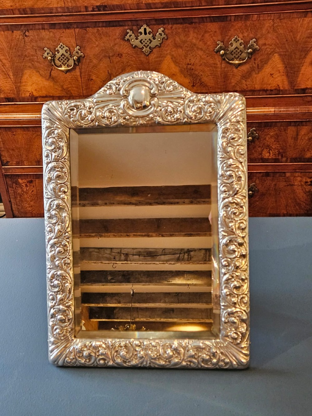 A Victorian easel-back mirror featuring an embossed silver frame decorated with scrollwork, acanthus leaves, and a central cresting cartouche around a bevelled glass plate. View VI