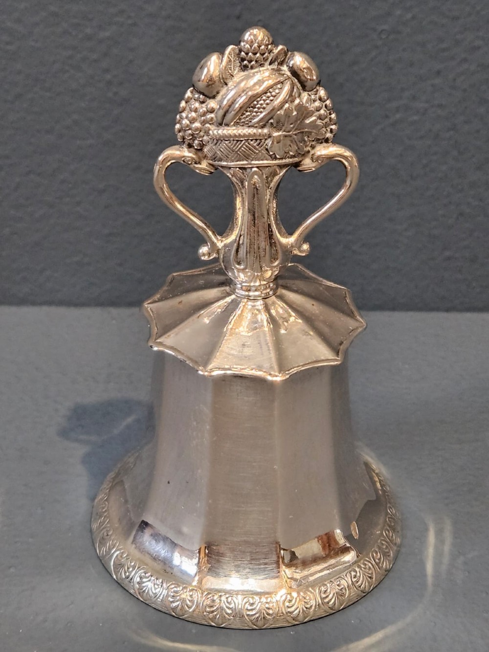 A Victorian silver table bell featuring a fruit basket finial, dual handles, a faceted body, and an embossed rim.