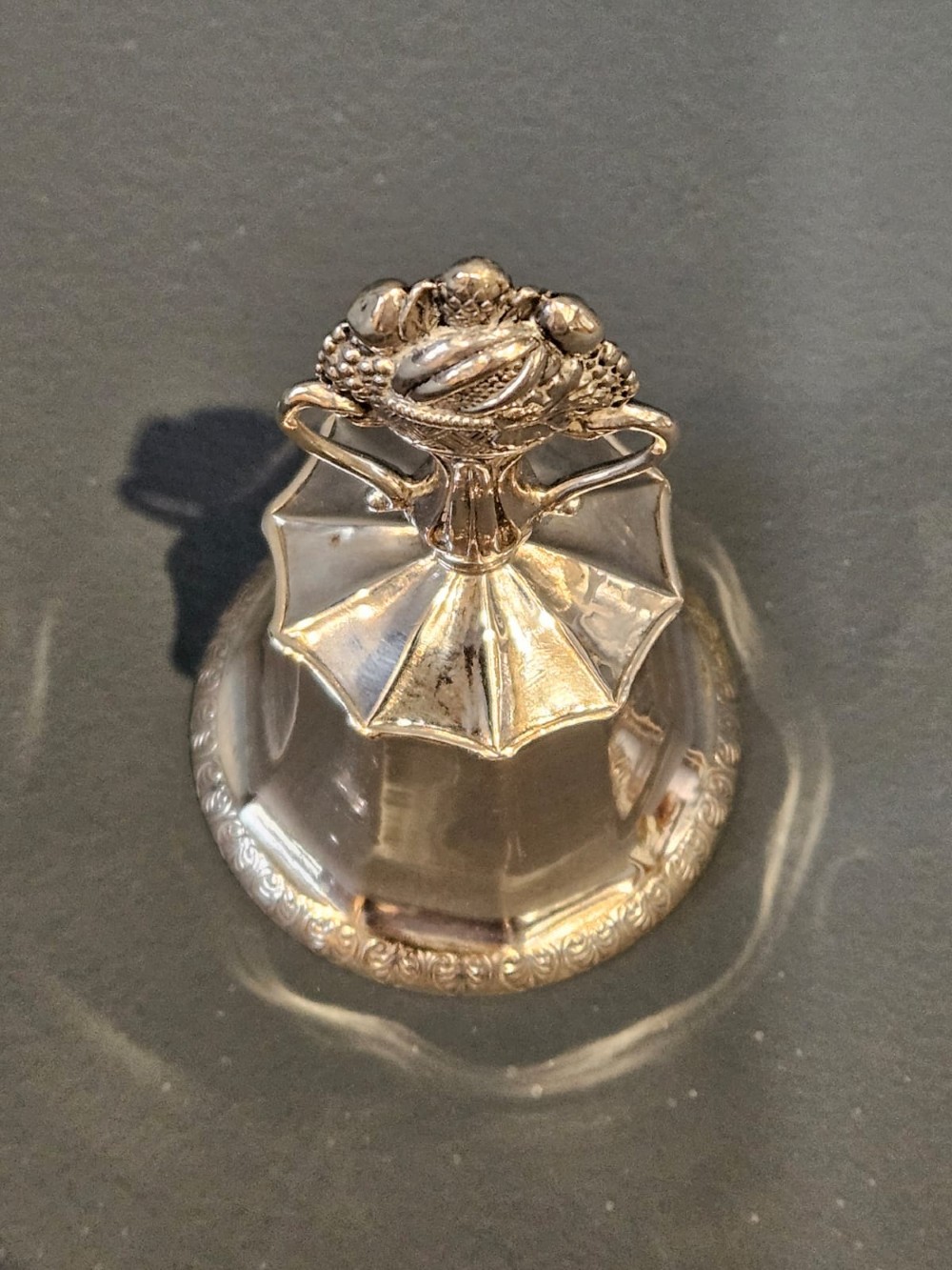 A Victorian silver table bell featuring a fruit basket finial, dual handles, a faceted body, and an embossed rim. View II